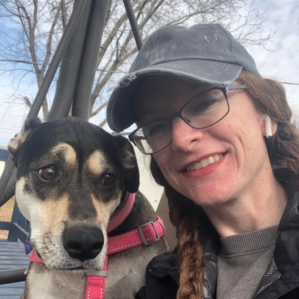 Image of author in gray cap and shirt in front of a tree, sitting beside her trusty dog.